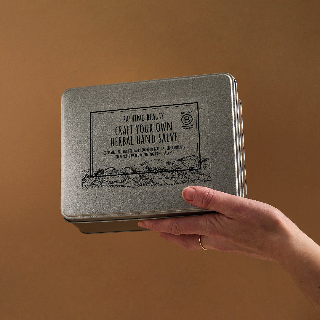 Hand holding a metal tin labeled 'Craft Your Own Herbal Hand Salve' against a brown background