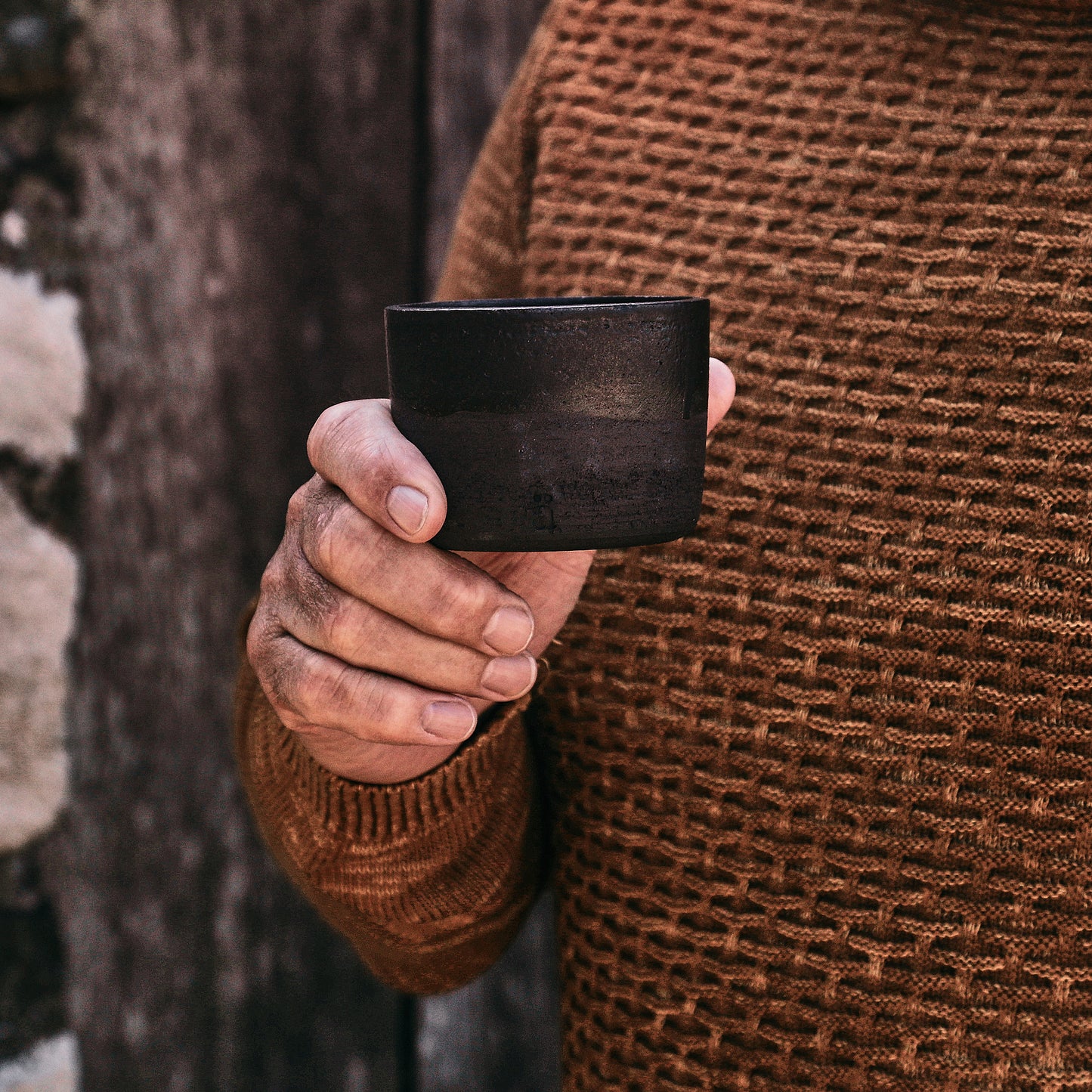 Person wearing a brown sweater holding a black wooden cup against a natural background