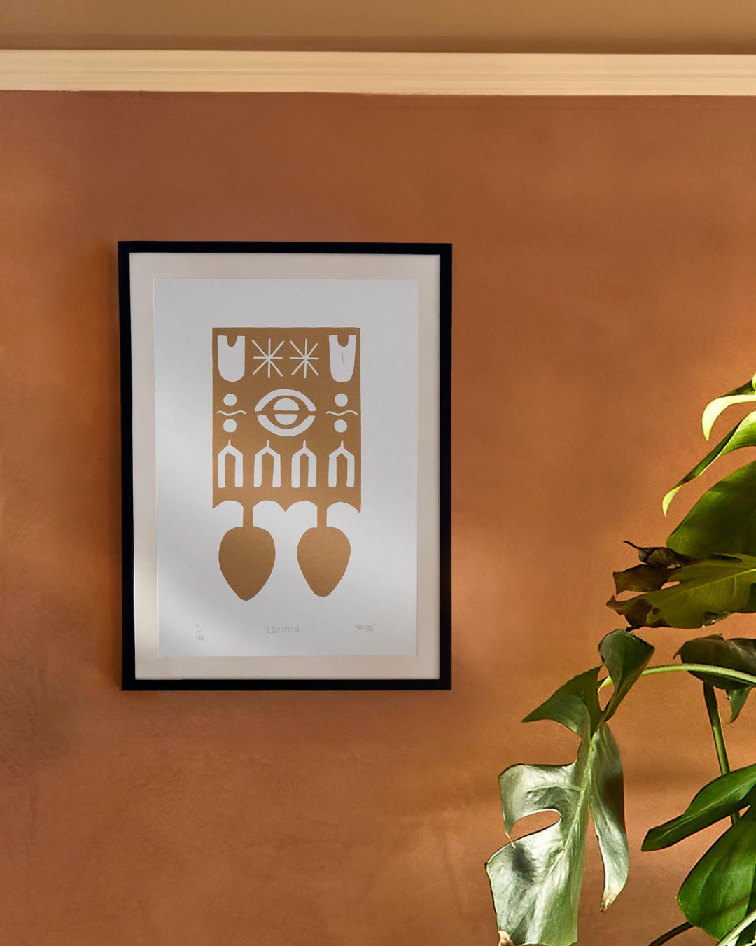 Framed artwork with welsh love spoon design on a orange wall with a plant in the foreground